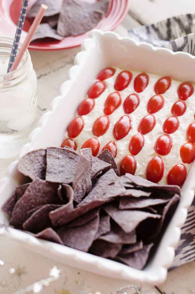 Patriotic Flag Parmesan Garlic Chip Dip is a festive appetizer recipe perfect for a holiday party with red, white and blue colors! A creamy Parmesan garlic dip is topped with cherry tomatoes and served with blue corn tortilla chips for a delicious dish sure to be a crowd pleaser.